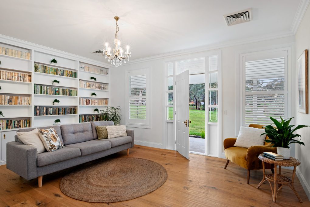 A charming sitting room and home library with warm natural hardwood timber flooring, styled with a grey linen sofa, a round jute rug, a mustard velvet armchair, and a floor-to-ceiling built-in bookshelf, all bathed in garden light streaming through open white French doors beneath an elegant crystal chandelier.