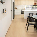 Light oak wood flooring in a modern, minimalist kitchen and dining area with white cabinetry, a white island, and black chairs.