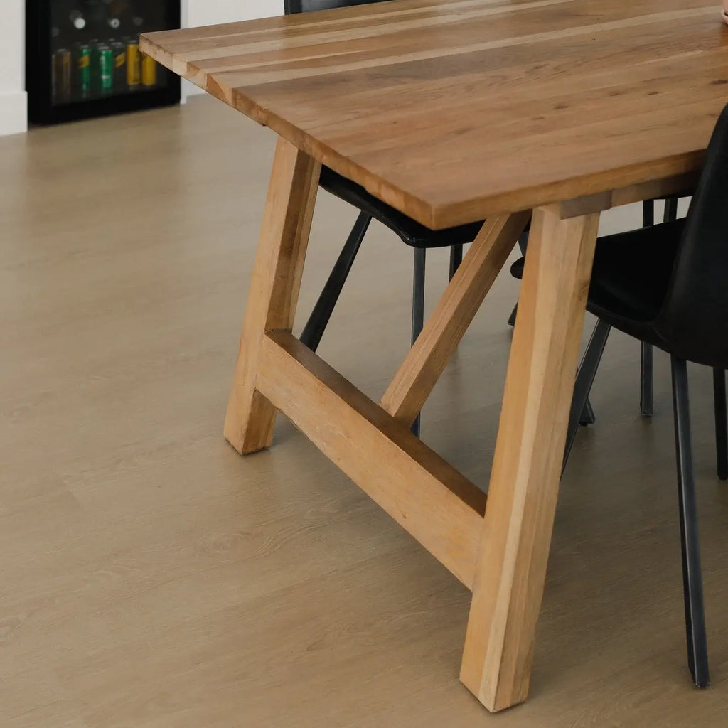 A close-up of a light natural wood dining table with a trestle-style leg base, shown alongside black chairs on a light wood floor.