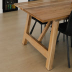 A close-up of a light natural wood dining table with a trestle-style leg base, shown alongside black chairs on a light wood floor.