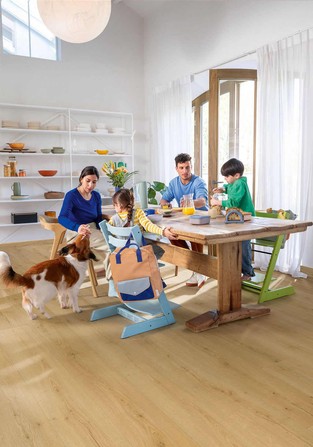 Family with children and a dog gathered around a wooden dining table in a bright room.