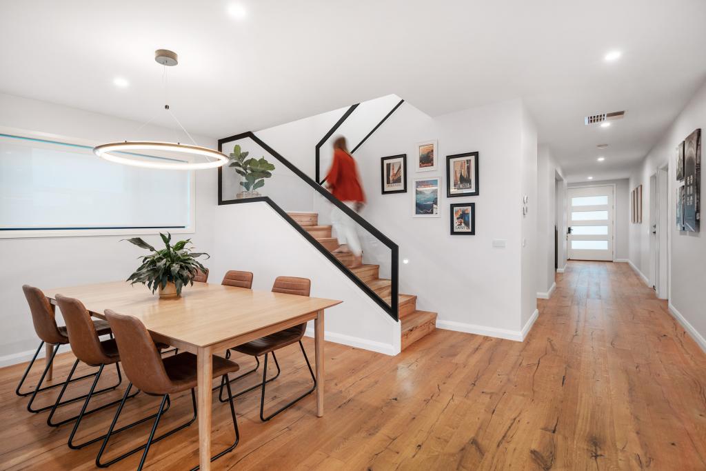 A wide-angle view of a bright, modern open-plan home featuring stunning Arcadian Oak hardwood timber flooring that flows from the dining area through to the hallway, anchored by a natural oak dining table, tan leather chairs, a circular pendant light, and a bold black steel staircase with matching timber treads.
