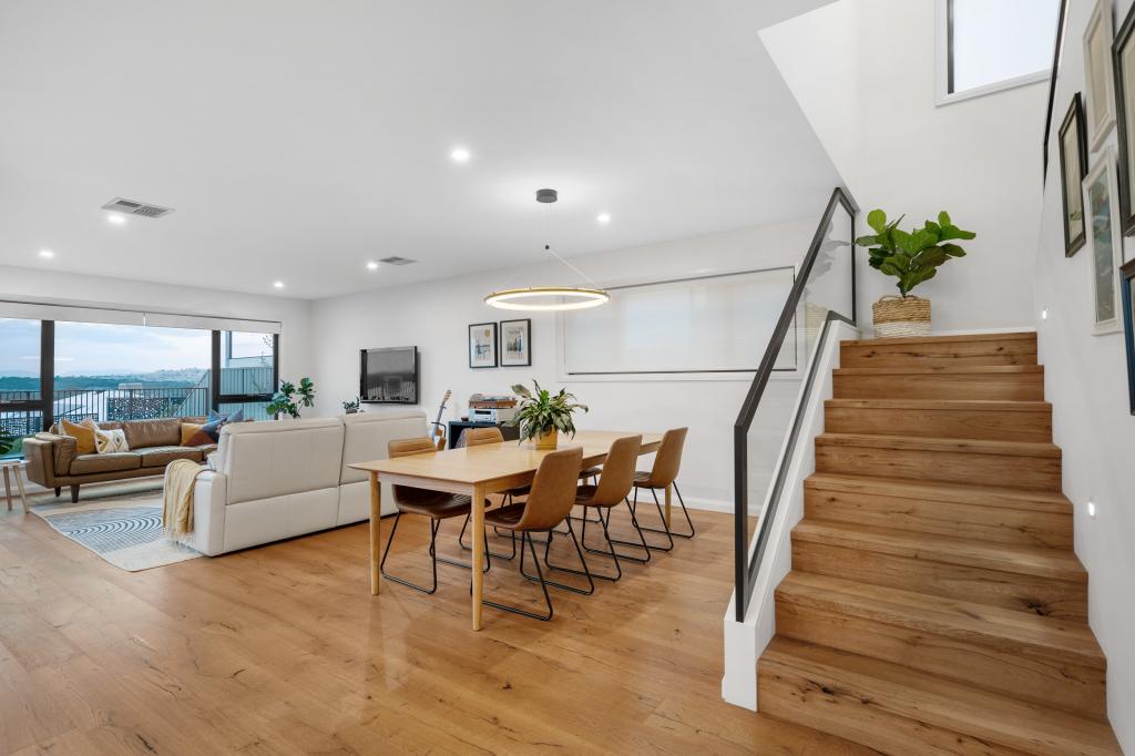 A spacious open-plan living and dining room with warm Arcadian Oak hardwood timber flooring flowing seamlessly from the staircase through to a stunning view of the cityscape beyond, styled with a natural oak dining table, tan leather chairs, a circular pendant light, and a relaxed neutral lounge setting.