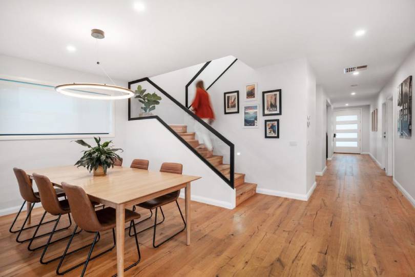 A bright, modern open-plan dining room featuring warm Australian hardwood timber flooring that flows seamlessly into the hallway, paired with a natural oak dining table, tan leather chairs, and a striking staircase with black steel balustrades.