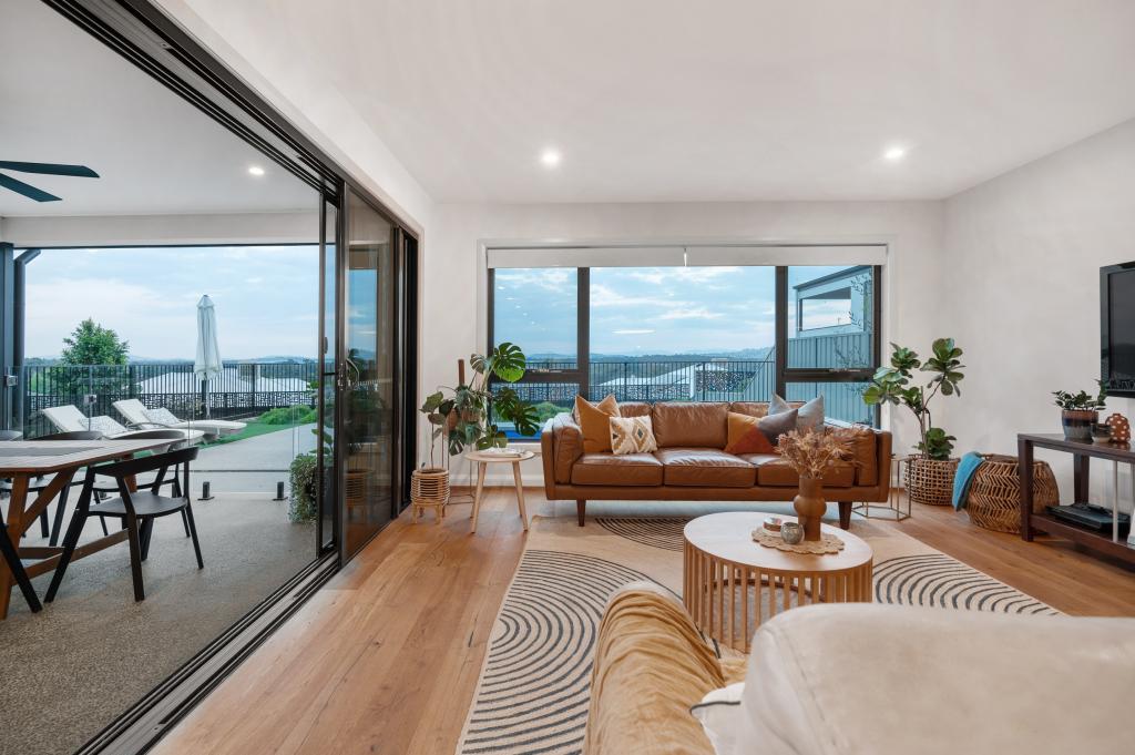 A breathtaking open-plan living room with warm Arcadian Oak hardwood timber flooring extending out toward full-height sliding glass doors that open to an alfresco entertaining area, framed by sweeping panoramic views, tan leather sofas, lush indoor plants, and earthy natural textures throughout.