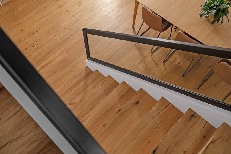 A stunning bird's-eye view of natural oak timber stair treads with visible character grain and knots, flowing down to matching hardwood timber flooring below, framed by sleek black steel balustrades and white risers for a modern, timeless finish.
