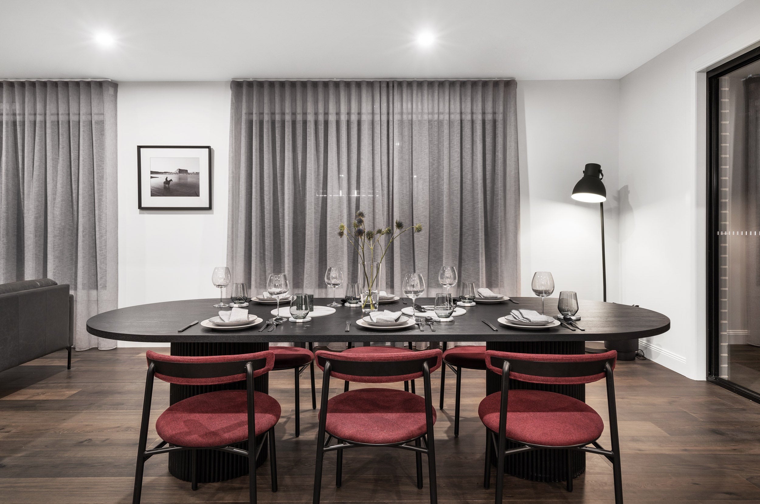 An elegant formal dining room with rich ash grey engineered timber flooring setting the tone for a sophisticated interior, featuring a beautifully set black oval fluted dining table, deep crimson upholstered chairs, floor-to-ceiling grey drapes, and a dramatic black floor lamp for a restaurant-quality dining experience at home.