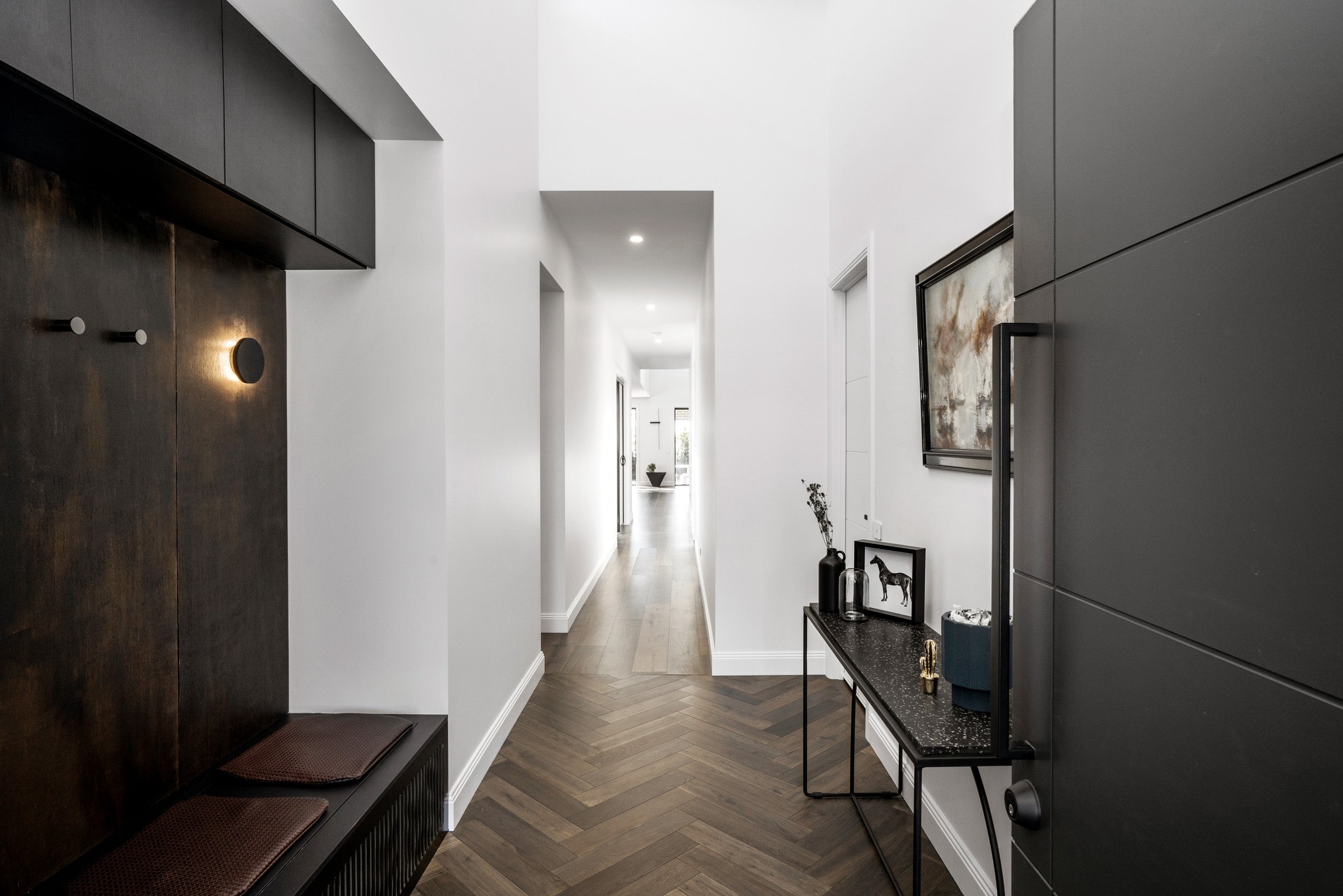 A dramatic home entrance featuring dark ash grey herringbone parquetry timber flooring that transitions seamlessly into straight-laid hardwood flooring down a long, bright hallway, styled with sleek grey cabinetry, an oxidised metal feature wall, a terrazzo console table, and moody ambient wall lighting.