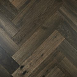 A close-up timber flooring sample of Ash Grey engineered oak laid in a classic herringbone parquetry pattern, showcasing its deep, smoky charcoal tones, subtle natural grain variation, and rich character that adds timeless elegance and sophistication to any interior space.
