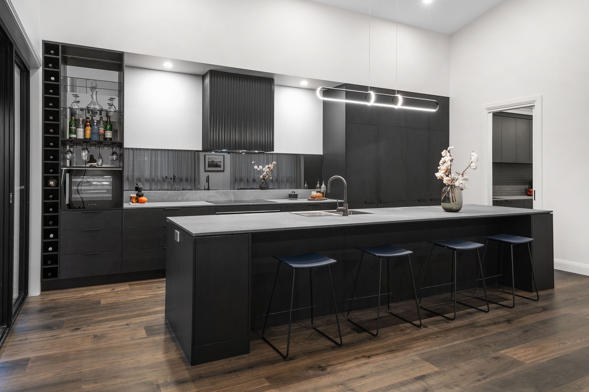A show-stopping entertainer's kitchen with rich dark ash grey engineered timber flooring anchoring an all-black kitchen featuring a lengthy stone-topped island bench, navy bar stools, a built-in wine rack, glass bar cabinet, and a statement tubular pendant light for a truly luxurious finish.