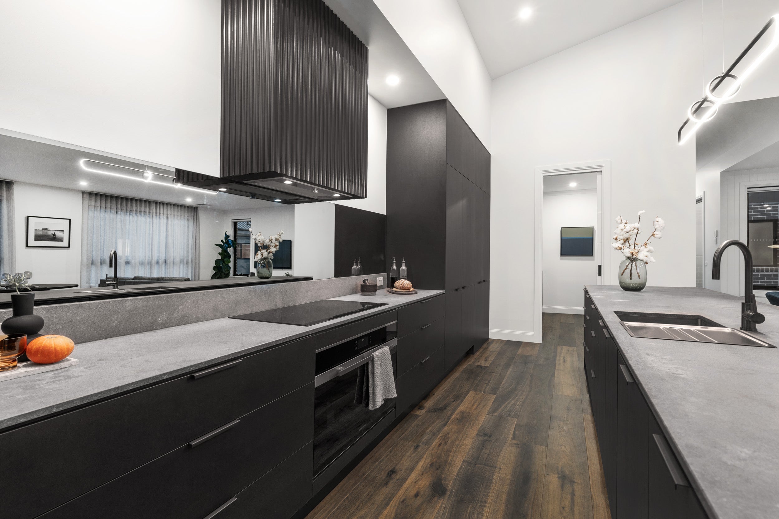A luxurious chef's kitchen featuring deep, dark ash grey engineered timber flooring that perfectly complements the matte black cabinetry, concrete-look stone benchtops, dramatic fluted rangehood, and mirror splashback for a bold, magazine-worthy contemporary interior.