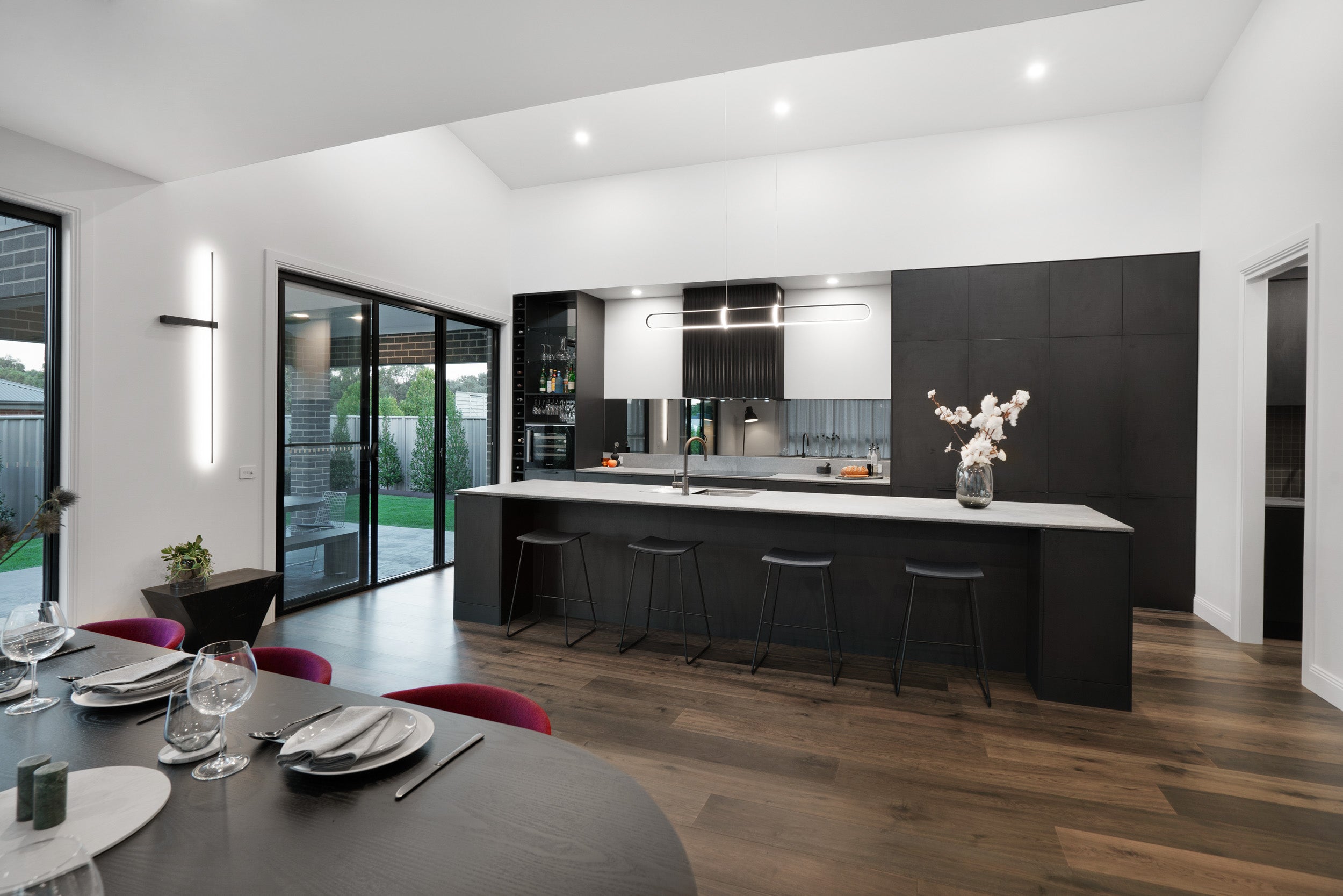 A dramatic high-end kitchen and dining space where deep ash grey engineered timber flooring grounds a striking all-black kitchen with a long stone island bench, sleek bar stools, and a built-in wine wall, all flowing seamlessly to a lush alfresco entertaining area through full-height sliding glass doors.