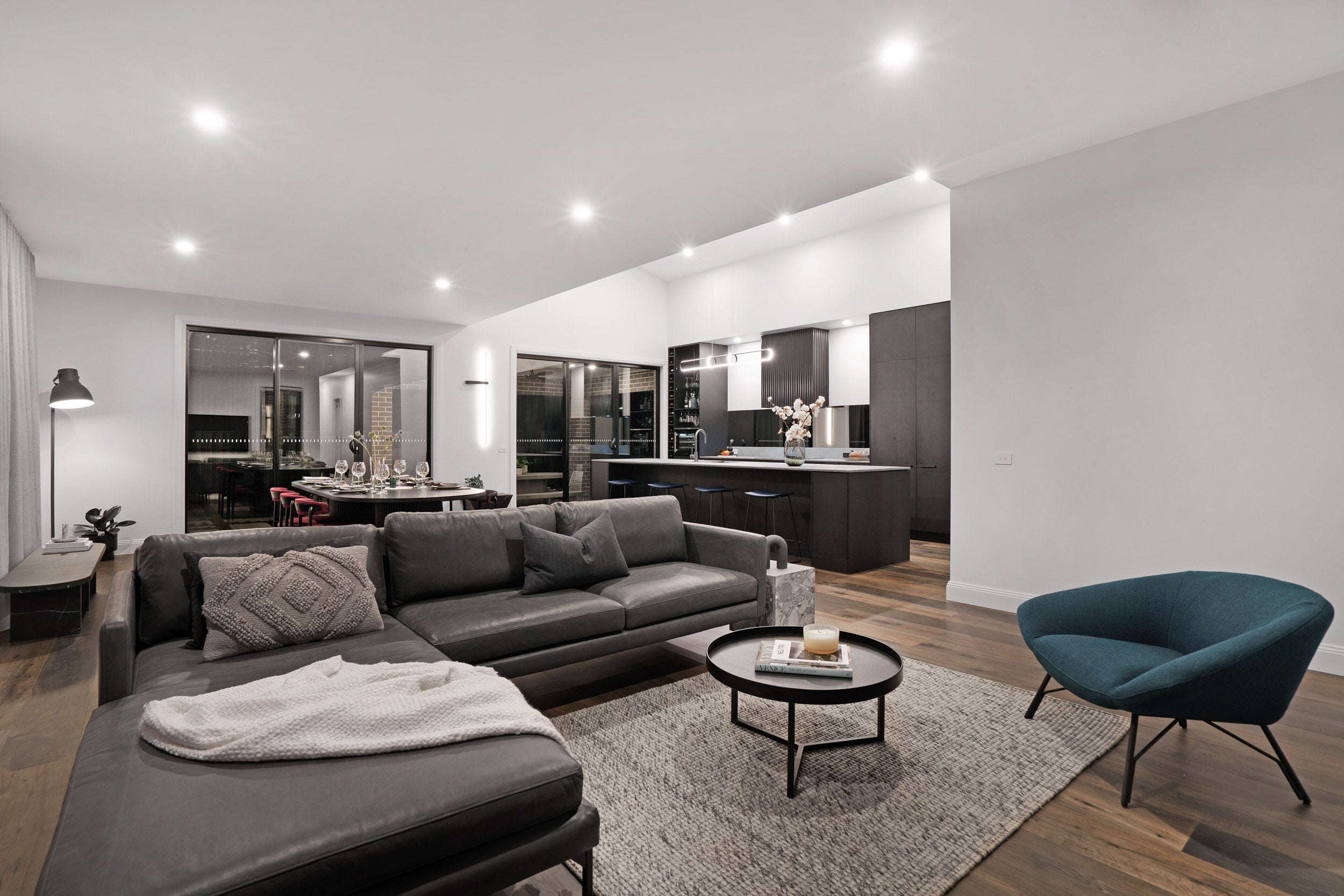 A sophisticated open-plan living space featuring rich ash grey engineered timber flooring that anchors the dramatic interior, styled with a charcoal leather sectional sofa, teal accent chair, dark cabinetry kitchen, and a glimpse of an elegantly set dining room beyond the glass doors.