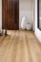 Light oak wood flooring shown in a modern interior hallway with a dark wood door, white walls, decorative vases, and a woven basket.