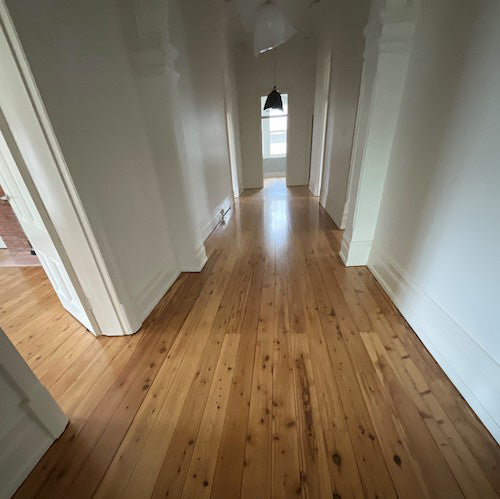A long, elegant hallway in a classic Australian federation home featuring freshly polished Cypress Pine timber floorboards with charming natural knots and warm golden tones, stretching toward a light-filled entrance and framed by crisp white architraves and high ceilings.