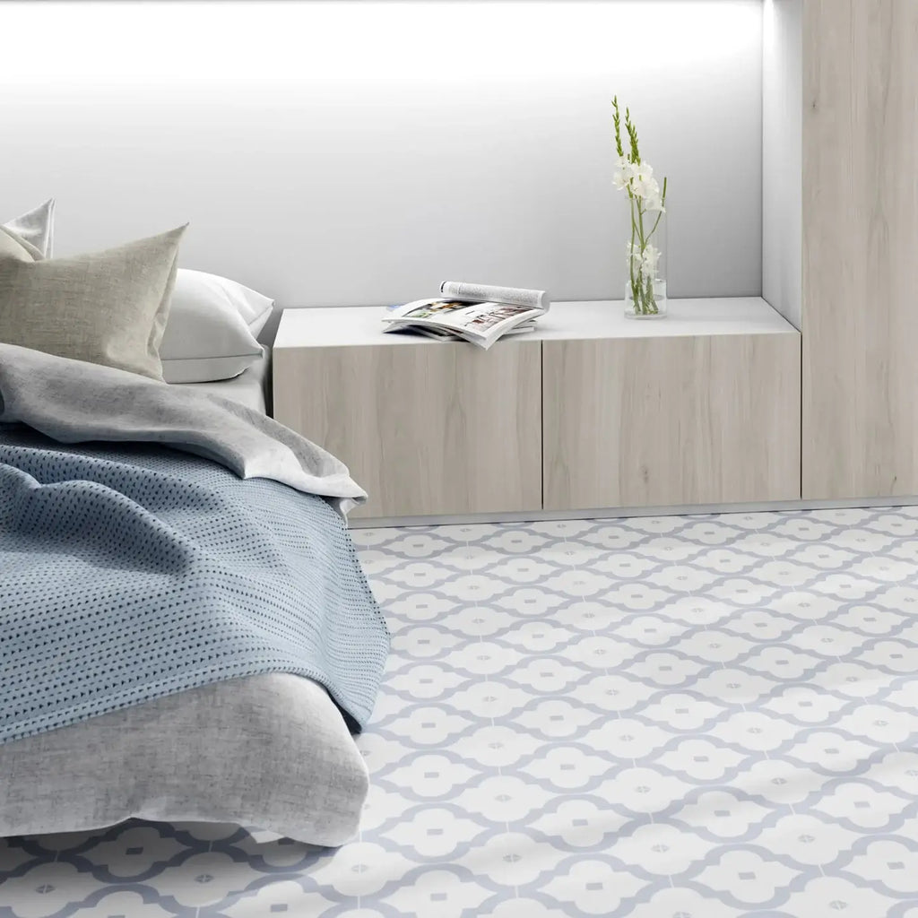 A modern bedroom scene showing a bed with gray and blue bedding, a light wood nightstand with a vase of flowers and an open magazine, and a light patterned floor.