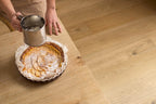 Person dusting an apple pie with flour using a sifter on a wooden table.