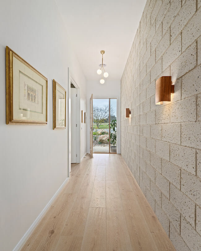 A stunning contemporary hallway featuring pale white oak engineered timber flooring that draws the eye toward a light-filled garden view, beautifully contrasted against a textured limestone feature wall, warm timber wall sconces, and elegant gold-framed artwork.