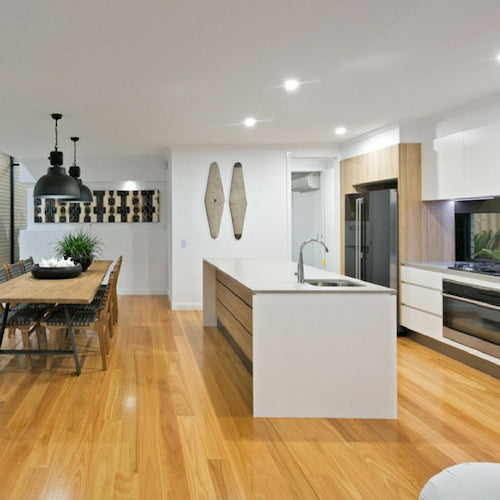 A bright, contemporary open-plan kitchen and dining area showcasing warm Blackbutt hardwood timber flooring that flows seamlessly throughout the space, complemented by white stone benchtops, timber veneer cabinetry, black pendant lights, and a rustic dining setting.
