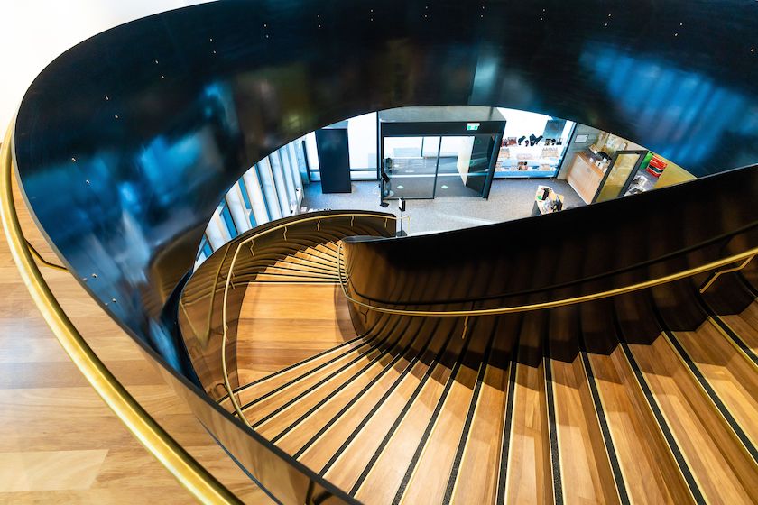 A breathtaking aerial view of a world-class spiral staircase featuring warm hardwood timber flooring and matching timber stair treads elegantly framed by a sweeping black steel structure with brass handrails, showcasing the extraordinary versatility and enduring beauty of commercial timber flooring in a stunning public building interior.