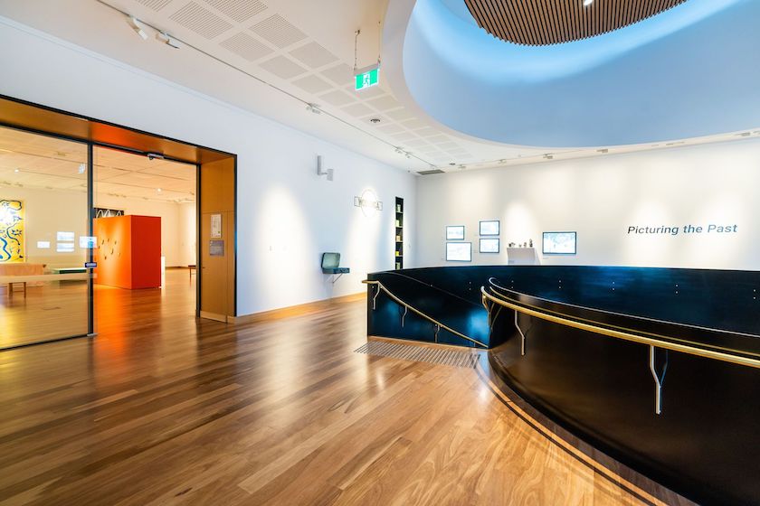 The upper gallery level of the Hyphen Wodonga Library and Gallery featuring beautifully polished hardwood timber flooring that flows through the light-filled exhibition space, wrapping elegantly around the dramatic black and brass spiral staircase opening and leading through to a vibrant gallery beyond.