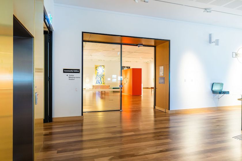 Warm, richly polished hardwood timber flooring flows through the entrance to the Community Gallery at the Hyphen Wodonga Library and Gallery, framed by sleek black-edged glass doors and timber detailing, demonstrating the warmth and durability of commercial hardwood timber flooring in a vibrant public cultural space.