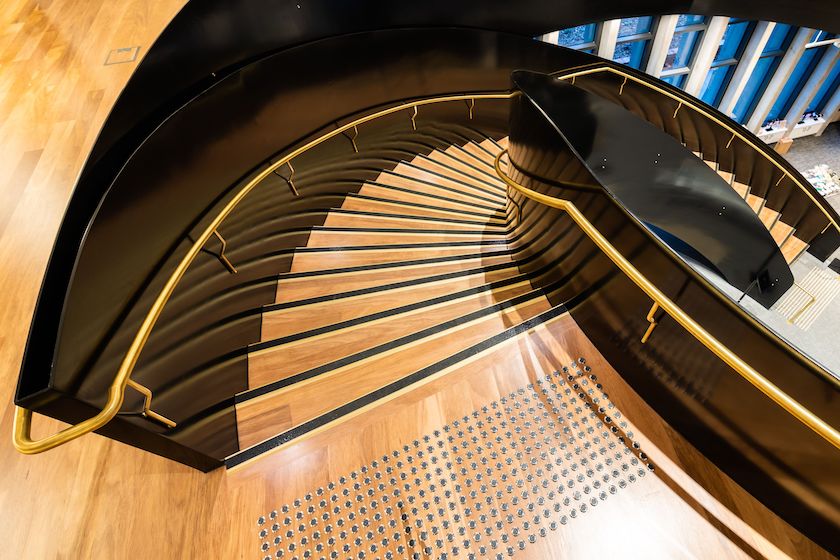 A stunning close-up overhead view of the iconic spiral staircase at the Hyphen Wodonga Library and Gallery, featuring warm hardwood timber stair treads and matching timber flooring elegantly contrasted against a dramatic black steel structure with gleaming brass handrails, a true masterpiece of commercial timber flooring craftsmanship.