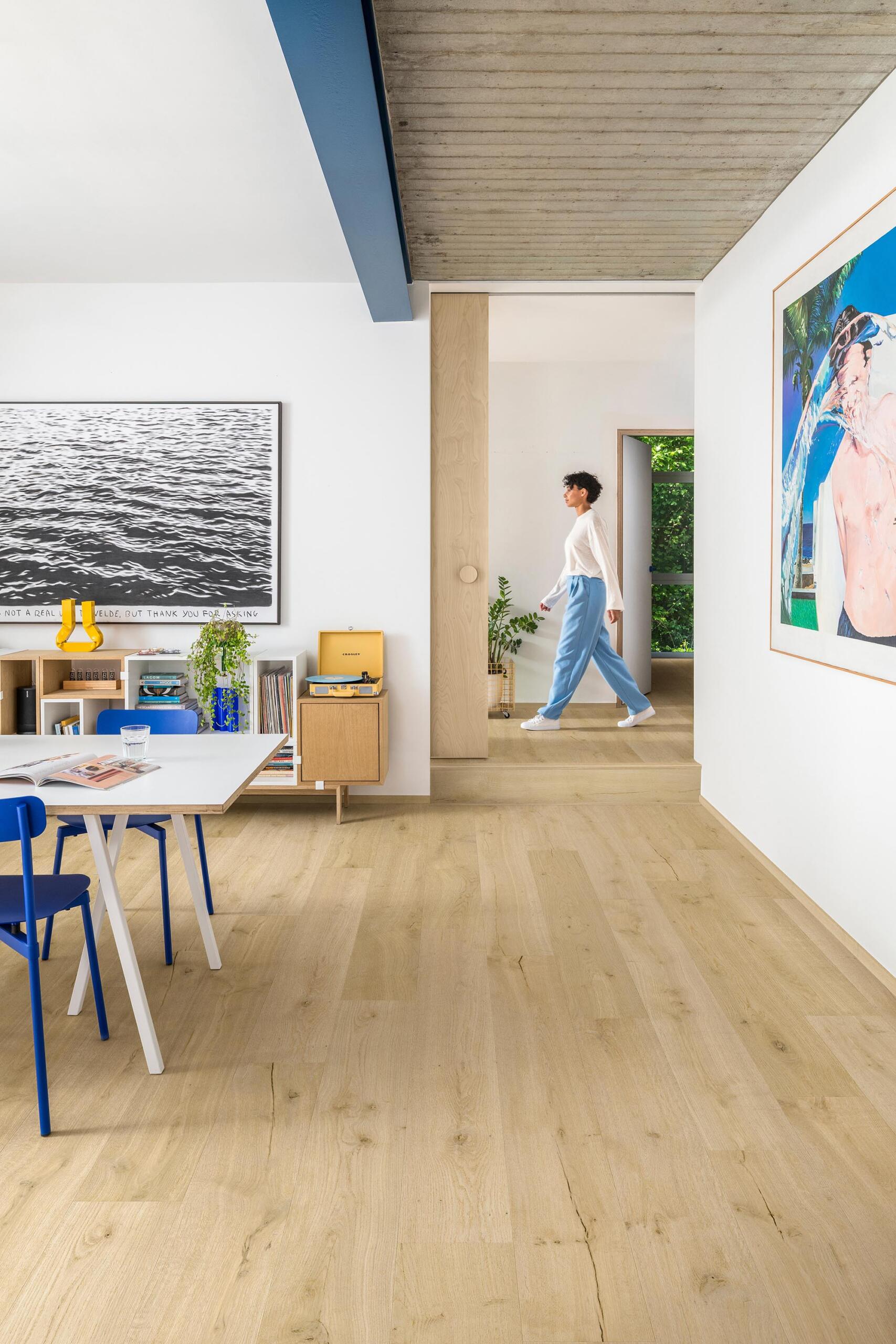 Light oak wood flooring in a modern, bright interior room with white walls, a dining table with blue chairs, and a person walking in the background hallway.