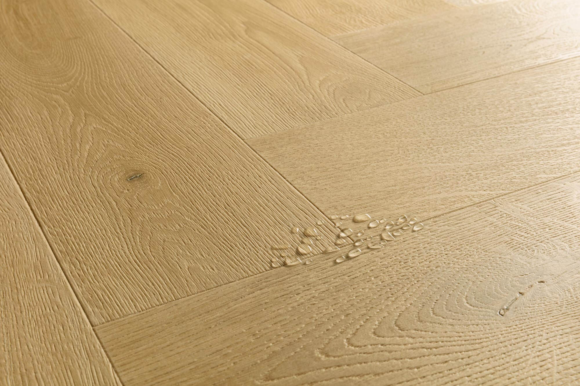 Light oak wood flooring planks arranged in a herringbone pattern, showing natural wood grain and a small cluster of water droplets on one plank.