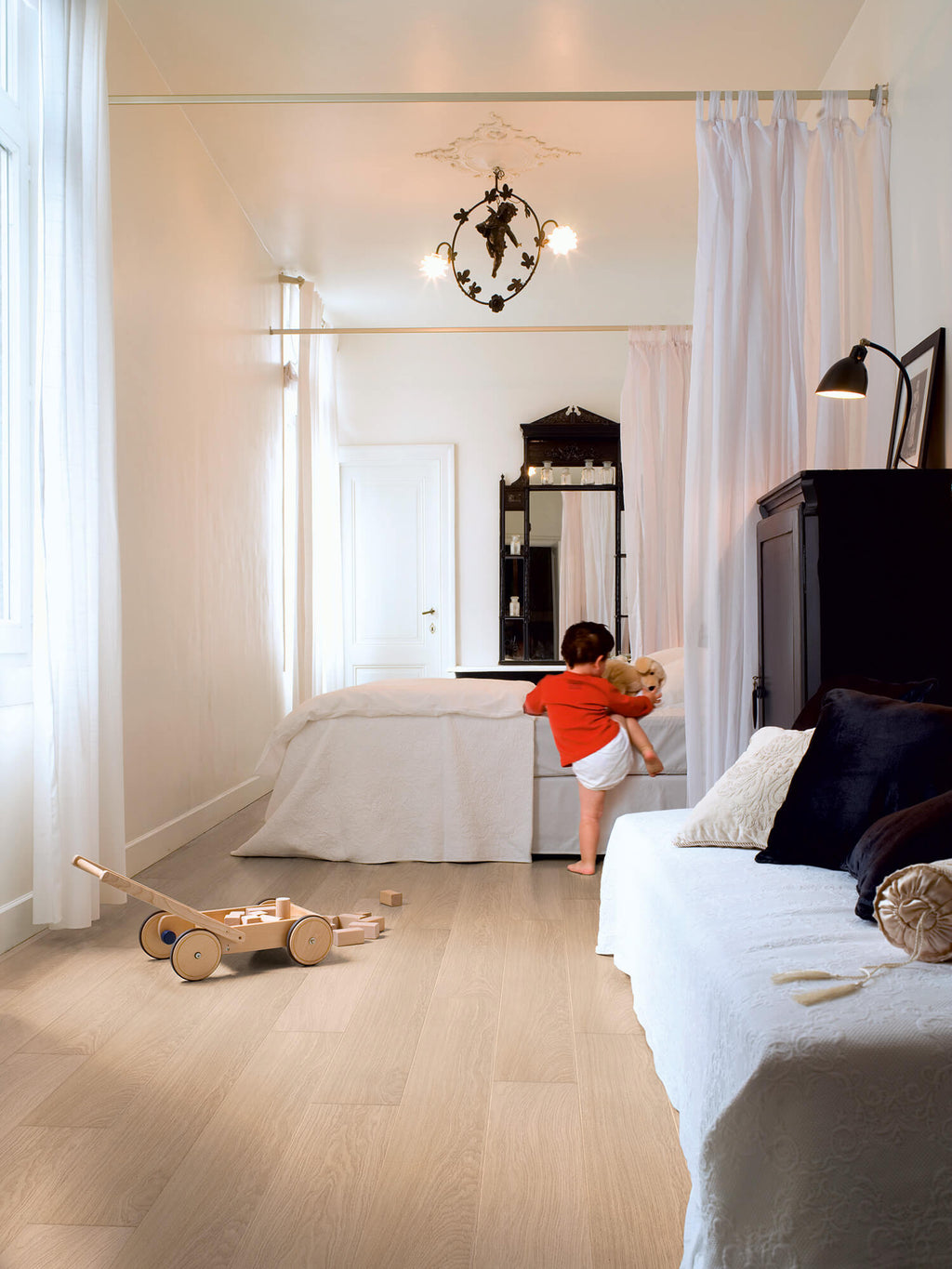 Child playing with a teddy bear in a bedroom with white curtains and wooden floor.