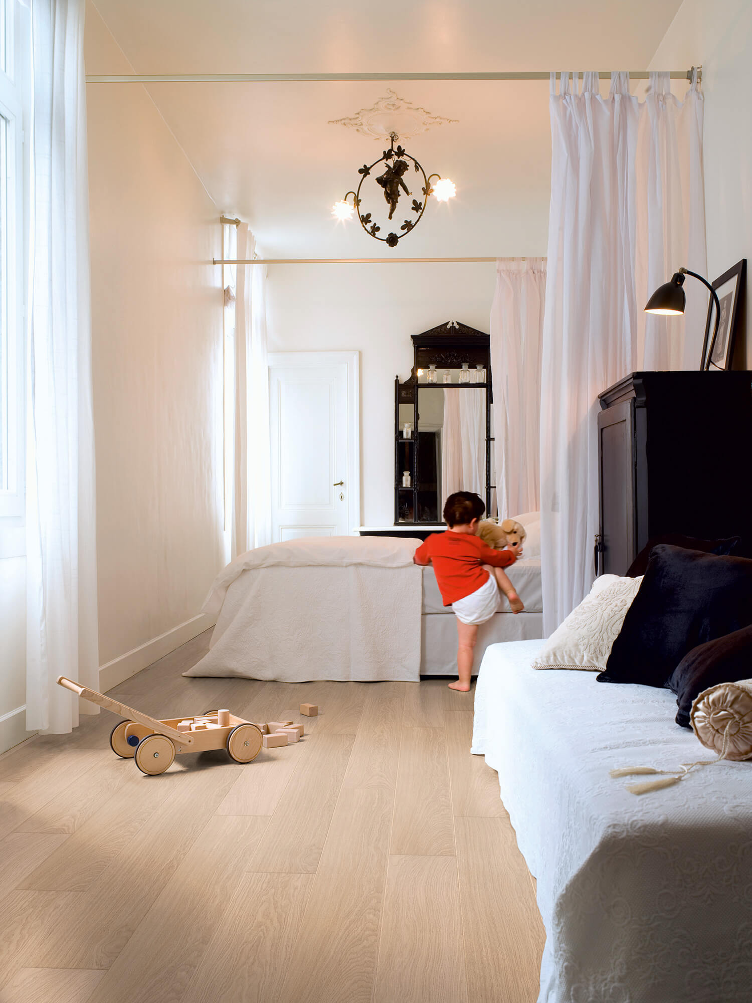 Child playing with a teddy bear in a bedroom with white curtains and wooden floor.