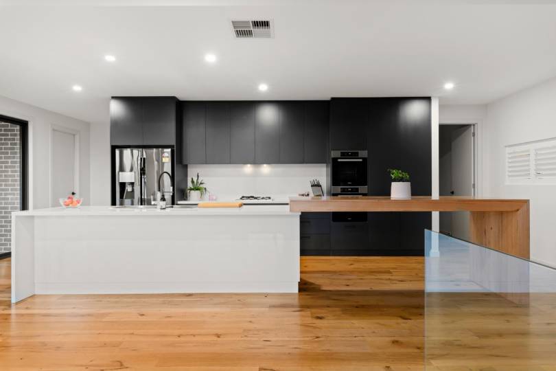 A sleek, contemporary kitchen with matte black cabinetry and a white stone island bench, elevated by warm natural timber flooring and a matching timber breakfast bar extension that adds a beautiful organic touch to the bold monochrome design.