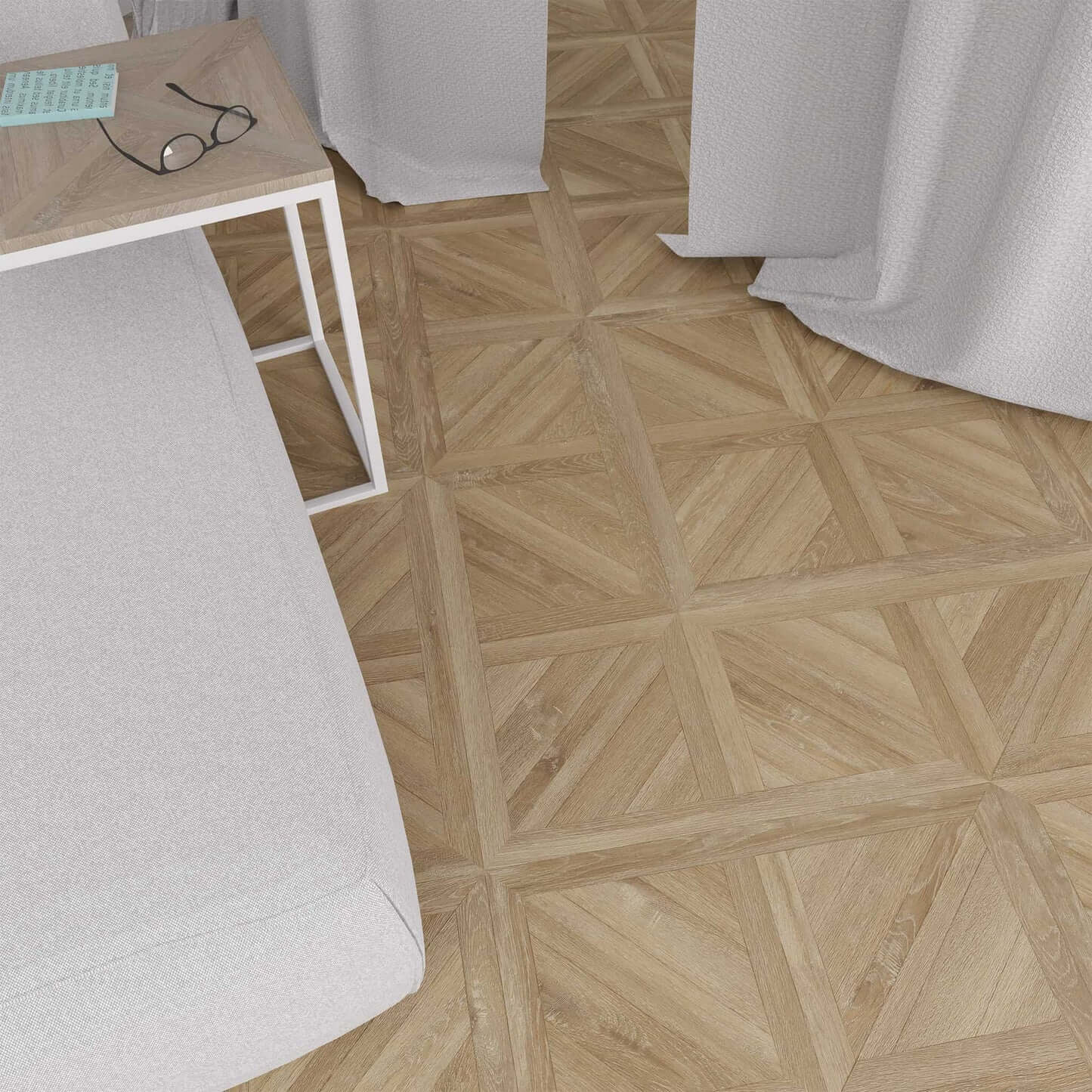 A top-down view of a light gray upholstered daybed or sofa next to a small light wood side table with a white metal frame. The room features a geometric parquet wood floor and white curtains.