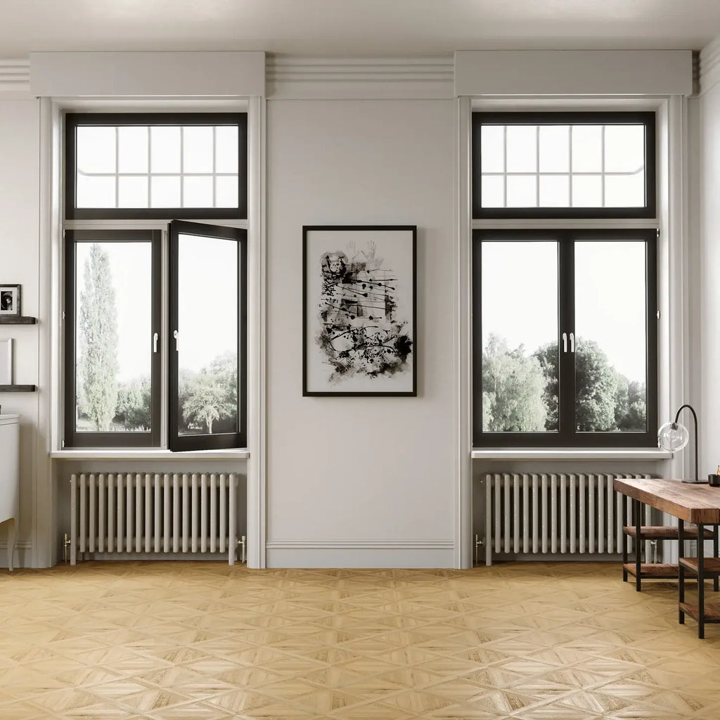 A modern interior room with two tall black-framed windows, a central framed black-and-white artwork on a white wall, radiators beneath the windows, a light wood floor, and part of a wooden desk on the right.