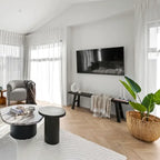 A modern, bright living room with light wood herringbone flooring, white walls, sheer white curtains, a gray armchair, a black wall-mounted TV, a black bench with a throw blanket, and a large potted green plant in a woven basket.