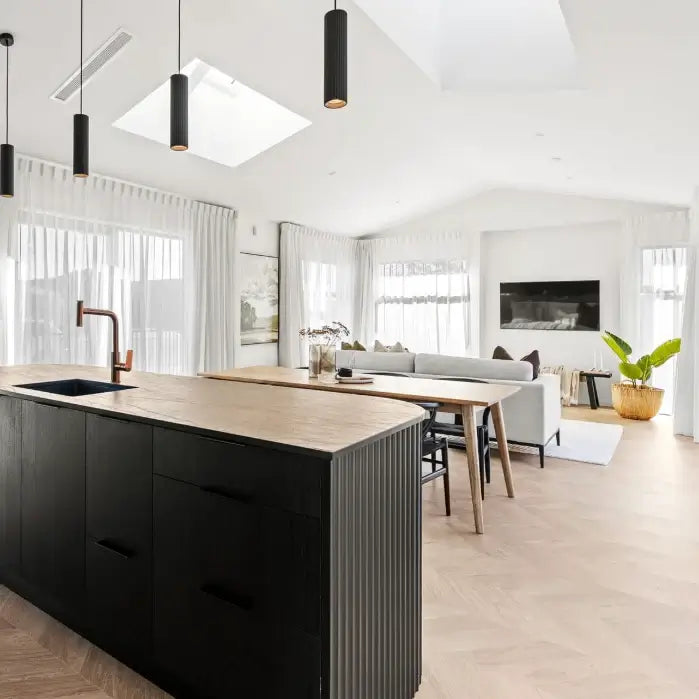 A modern kitchen island with a matte black ribbed cabinet body and a light wood countertop, set in a bright, contemporary open-plan living space with light wood floors and white walls.
