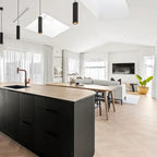 A modern kitchen island with a matte black ribbed cabinet body and a light wood countertop, set in a bright, contemporary open-plan living space with light wood floors and white walls.