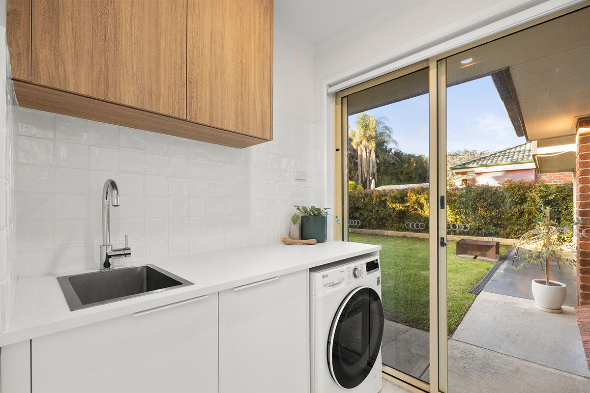 A fresh and functional laundry room with warm soft oak timber veneer overhead cabinetry, white stone benchtop, and glossy subway tile splashback, opening through sliding glass doors to a sunny, well-established garden — a practical space that carries the same natural warmth as the home's timber flooring throughout.