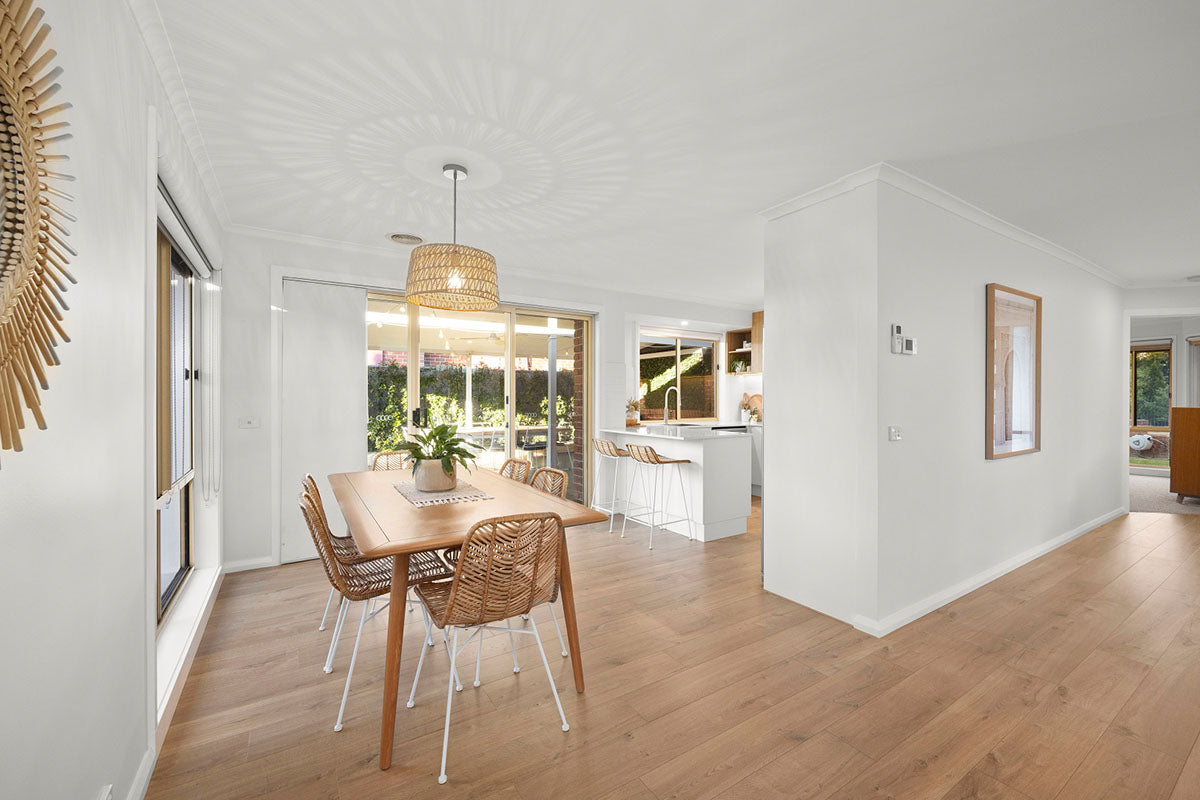 A bright and breezy open-plan dining and kitchen space with warm soft oak natural timber flooring flowing throughout, styled with a timber dining table, woven rattan chairs, a wicker pendant light, and rattan bar stools at a white kitchen island, all bathed in beautiful natural light from garden-facing sliding doors.
