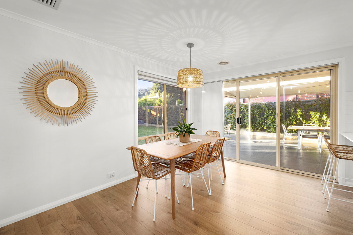 A sun-drenched dining room with warm soft oak natural timber flooring flowing through to a lush garden beyond full-width sliding glass doors, styled with a timber dining table, woven rattan chairs, a wicker pendant light, and a statement sunburst mirror for a relaxed, nature-inspired Australian home.