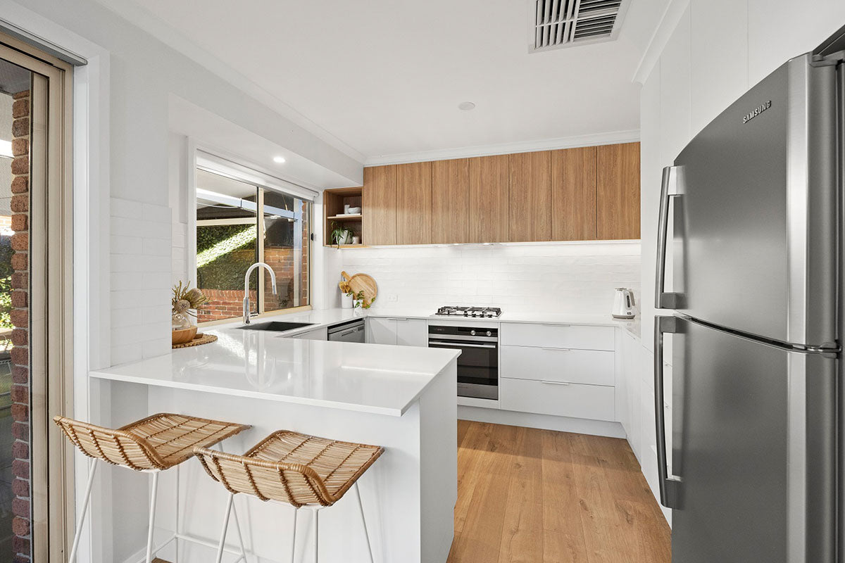 A light and airy family kitchen with warm soft oak natural timber flooring tying together the timber veneer overhead cabinetry and woven rattan bar stools, complemented by a white stone breakfast bar, crisp subway tile splashback, and a garden view through the window for a fresh, nature-inspired feel.