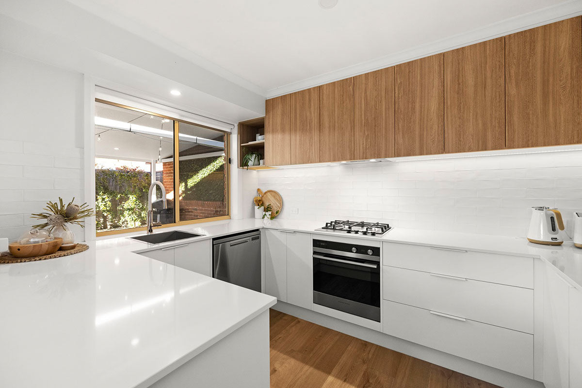 A clean and contemporary kitchen featuring warm soft oak natural timber flooring that beautifully echoes the rich timber veneer overhead cabinetry, paired with crisp white lower cabinets, a white stone benchtop, subway tile splashback, and a garden-facing window that fills the space with natural light.