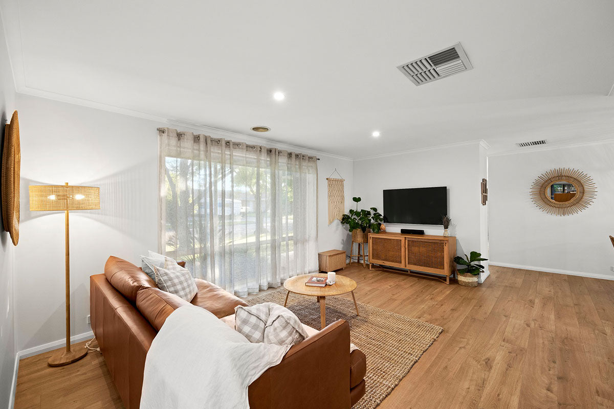 A cosy and relaxed Australian family living room with warm soft oak natural timber flooring, styled with a tan leather sofa, jute rug, rattan TV cabinet, woven floor lamp, macramé wall hanging, and a sunburst mirror that together create an effortlessly beautiful coastal boho feel.