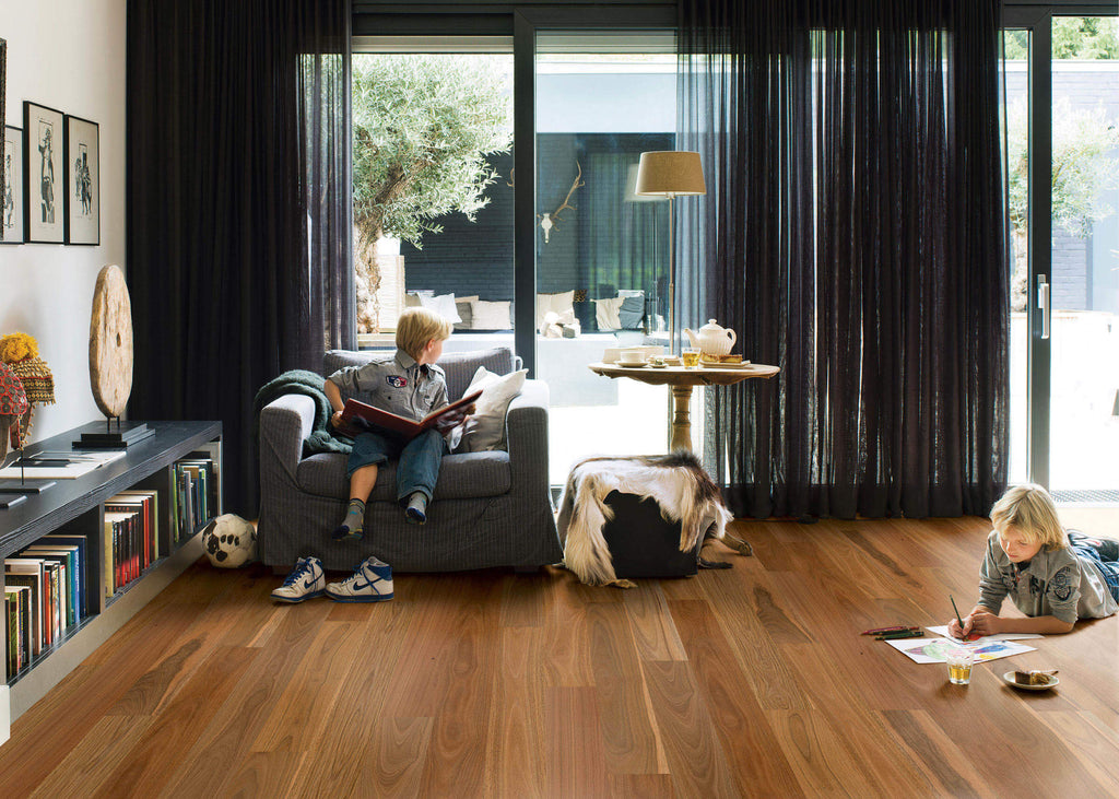 Two children in a modern living room with large windows, one reading a book on a sofa and the other at a small table.