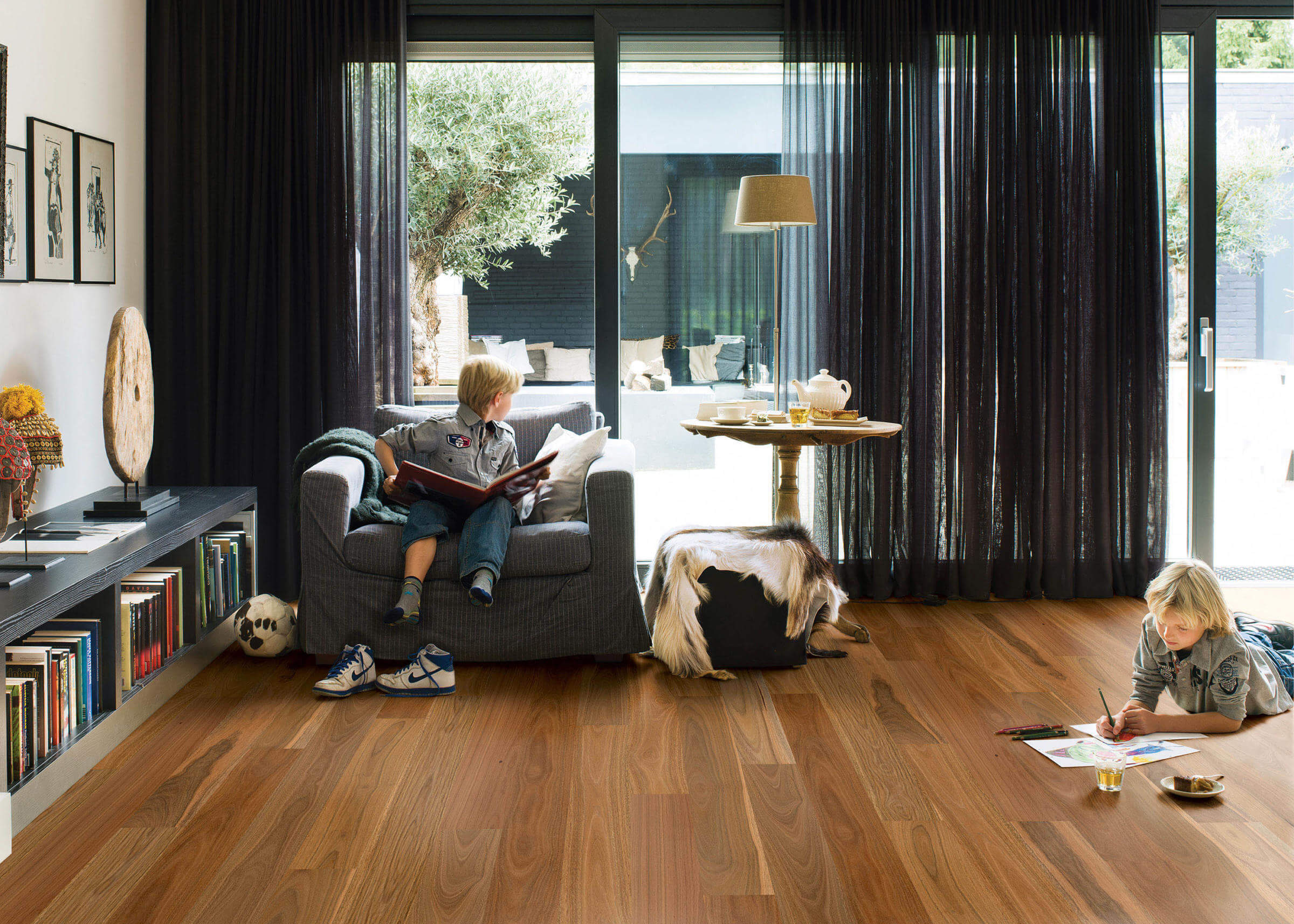 Two children in a modern living room with large windows, one reading a book on a sofa and the other at a small table.