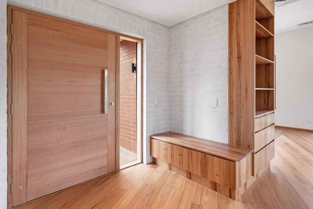 A beautifully crafted home entrance featuring warm Australian hardwood timber flooring that flows seamlessly beneath a stunning oversized solid timber pivot door, complemented by a custom-built hardwood timber joinery unit with open shelving, a built-in bench seat, and drawer storage set against a whitewashed brick wall.