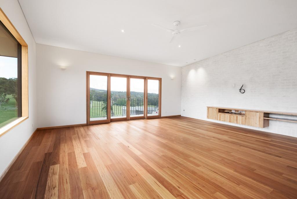 A spacious, light-filled living room in a contemporary rural home showcasing stunning Australian hardwood timber flooring with beautiful natural colour variation, framed by timber-framed French doors opening to sweeping countryside views, a whitewashed brick feature wall, and a floating timber TV cabinet.