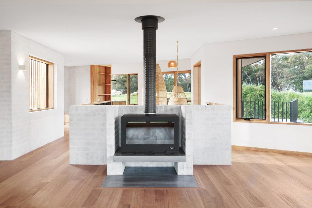 A striking open-plan rural home interior featuring warm Australian hardwood timber flooring flowing around a dramatic freestanding whitewashed brick fireplace with a black steel flue, framed by timber-framed windows with lush green countryside views and beautiful natural timber joinery and pendant lighting throughout.