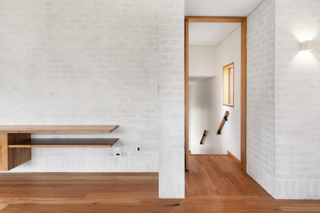 A serene and minimalist upper level hallway featuring warm Australian hardwood timber flooring with natural character grain, framed by whitewashed brick walls, a timber-framed doorway leading to a staircase with a sleek black timber handrail, and a floating hardwood timber shelf adding a simple, organic touch.