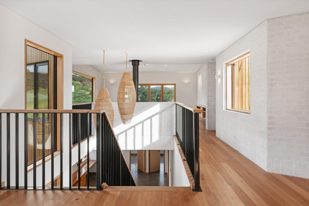 A light-filled upper landing in a contemporary rural home showcasing warm Australian hardwood timber flooring that flows through the open hallway, framed by black steel balustrades, timber-framed windows with lush green bush views, woven pendant lights, and a whitewashed brick feature wall.